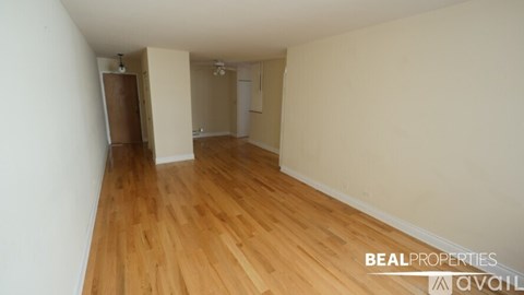 A room with wooden flooring and white walls, with a door on the left and a window on the right.