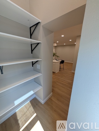 A hallway with a white wall and a wooden floor leading to a kitchen area.