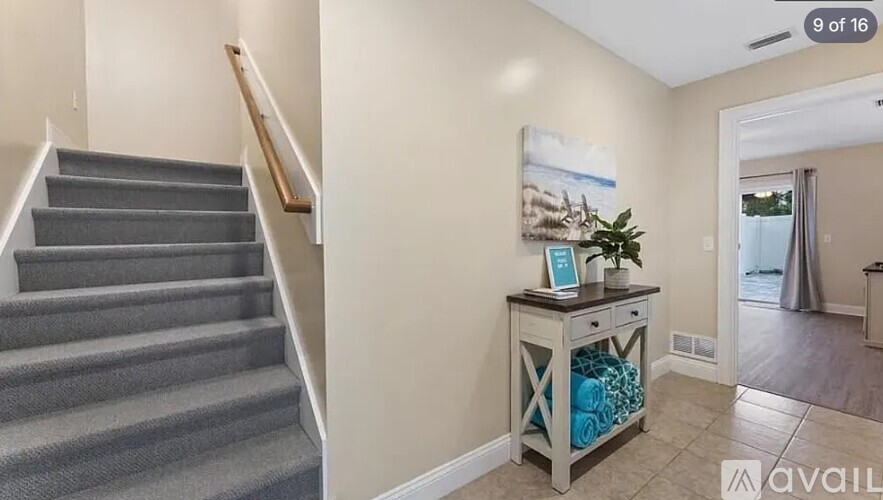A staircase with a grey carpeted runner and wooden handrail leads to an open space with a console table holding a plant and a painting.