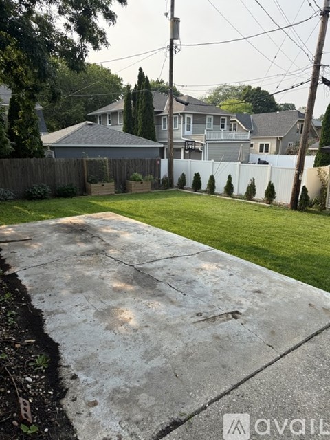 A concrete driveway leads to a house with a white fence and green lawn.