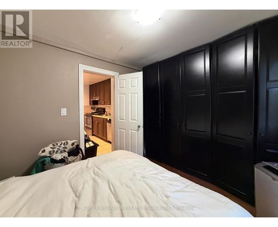 A bedroom with a bed in the foreground and a kitchen area visible through the open door.