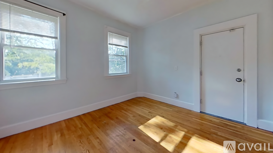 A room with wooden flooring and white walls, featuring a window with blinds, a door, and a small window on the wall.