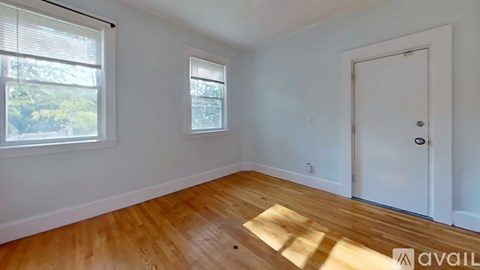 A room with wooden flooring and white walls, featuring a window with blinds, a door, and a small window on the wall.