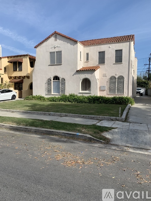 A white house with a red tile roof is situated on a street with a car parked in front.