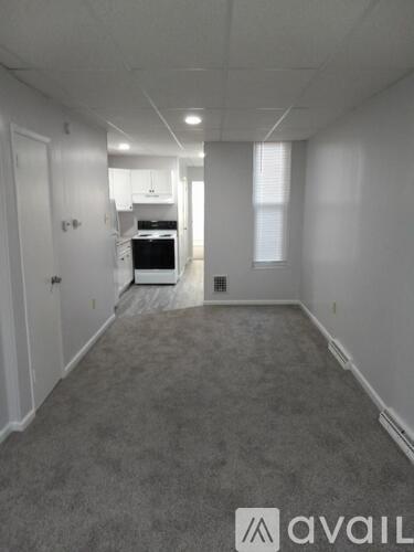 A room with a carpeted floor and a kitchen area in the background.