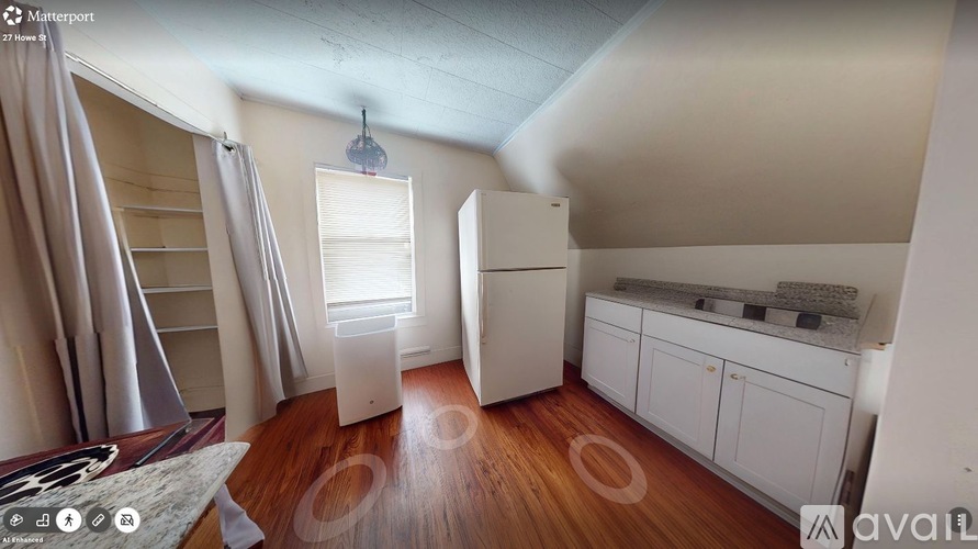 A room with wooden floors and white walls, featuring a white cabinet, a window with a curtain, and a hanging light fixture.