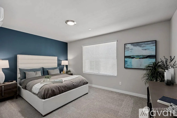 A bedroom with a large bed, a nightstand with a lamp, a desk with a plant, and a painting on the wall.