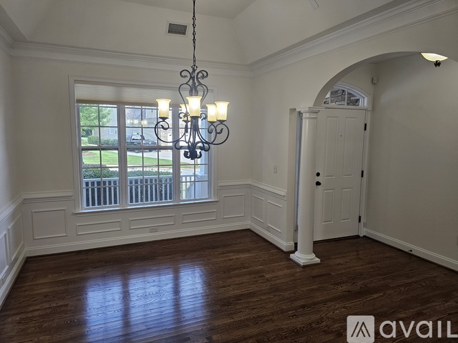 A room with a chandelier, a window with a view of a fence and trees, and a wooden floor.