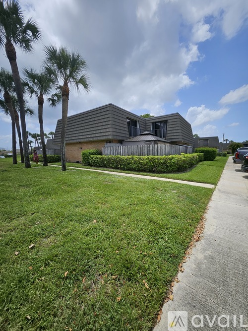 A modern building with a grey roof is surrounded by greenery and palm trees.