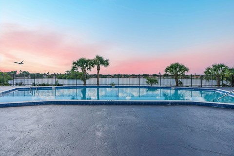 A swimming pool surrounded by a fence with palm trees and a plane flying in the sky.