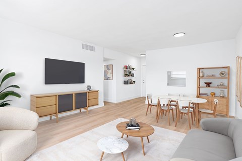 A living room with a grey sofa, a wooden coffee table, a white rug, a wooden cabinet, a flat screen TV, a dining table with chairs, and a potted plant.