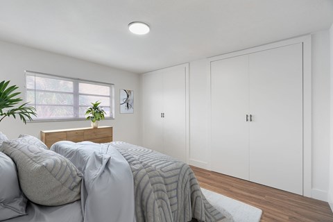 A bedroom with a bed covered in a grey blanket and a window with a plant on the windowsill.