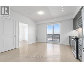 A well-lit, empty room with a kitchen area to the right and a door to the left.