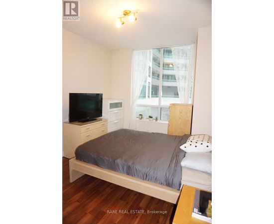 A bedroom with a bed, a television, and a window with white curtains.