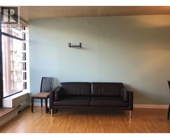 A black leather couch sits in a living room with a wooden floor and a wall mounted shelf.