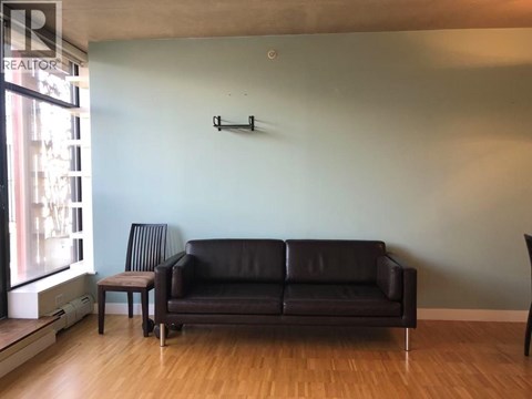 A black leather couch sits in a living room with a wooden floor and a wall mounted shelf.