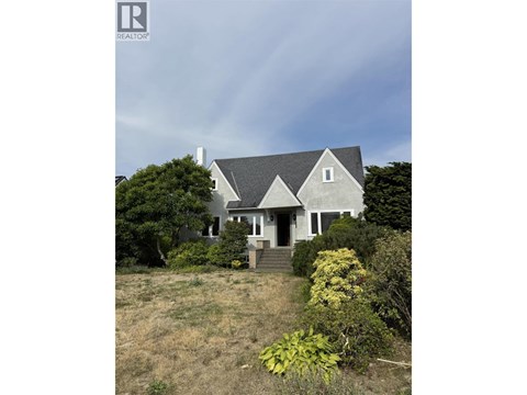 A house with a grey roof and white walls is surrounded by greenery.