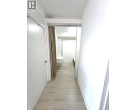 A hallway with a door on the left and a bedroom with a bed and a window on the right.