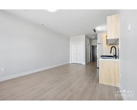 A spacious room with wooden flooring and white walls, featuring a kitchenette with a sink and cabinets.
