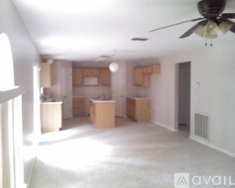 A living room with a ceiling fan and a kitchen area in the background.