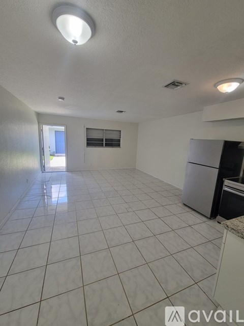 A spacious empty room with white tiled flooring and a kitchen area with a refrigerator.