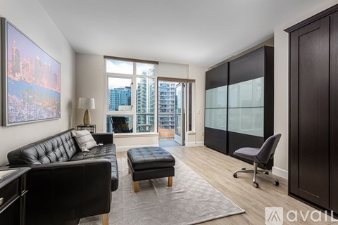 A living room with a black leather couch and ottoman, a grey rug, and a large window with a view of a city skyline.