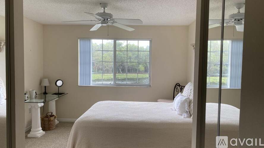 A bedroom with a bed, a nightstand, a lamp, a fan, and a window with a view of trees.