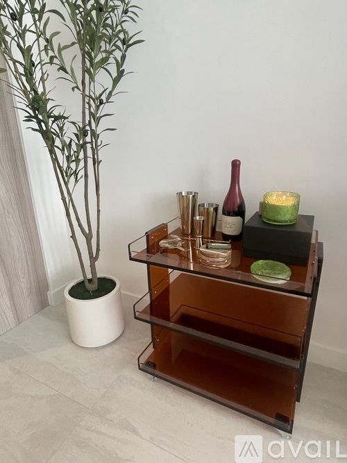 A small tree in a white pot sits next to a shelf with a wine bottle and candle on it.