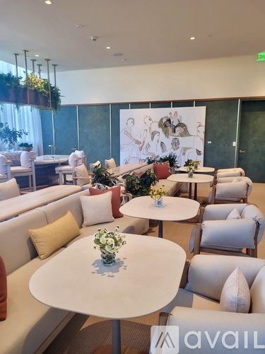 A white table with a vase of flowers on it sits in the middle of a room with white couches and a large painting in the background.