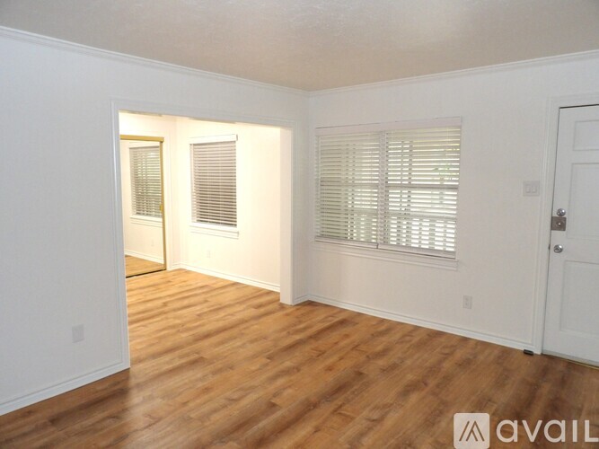 A room with wooden floors and white walls, with a door on the right and a window with blinds on the right wall.