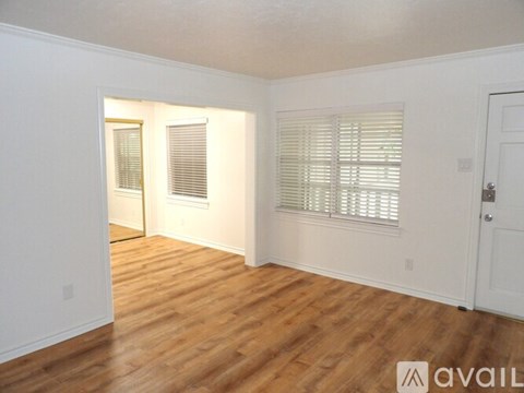 A room with wooden floors and white walls, with a door on the right and a window with blinds on the right wall.