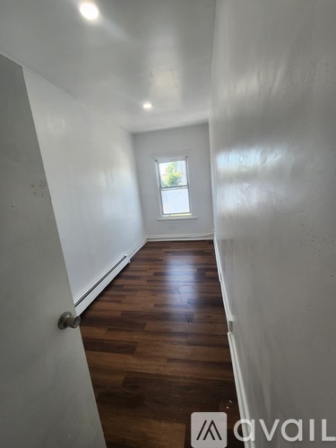 A hallway with wooden floors and white walls leading to a window.