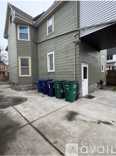 A house with a grey siding and a white door is shown with three green bins in front.