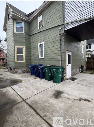 A house with a grey siding and a white door is shown with three green bins in front.