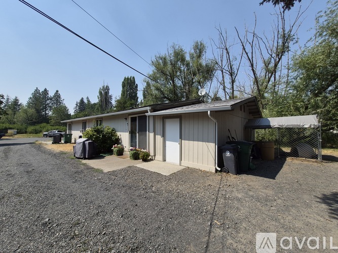 A gravel driveway leads to a white building with a metal roof and a fence.