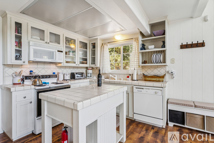 A kitchen with white cabinets and appliances, a black countertop, and a window with white curtains.