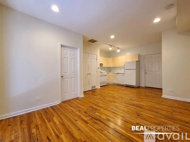 A room with wooden floors and white walls, with a refrigerator and cabinets in the background.