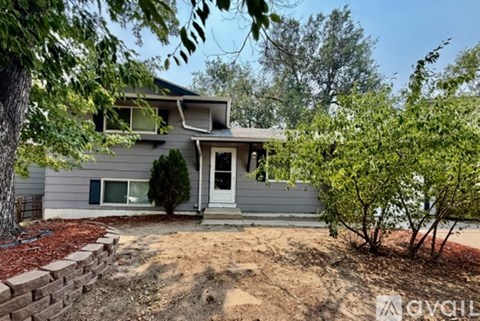 A house with a grey exterior and a white door is surrounded by trees and shrubs.