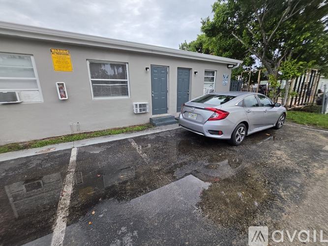 A grey car is parked in a paved lot in front of a grey building with a yellow sign on it.