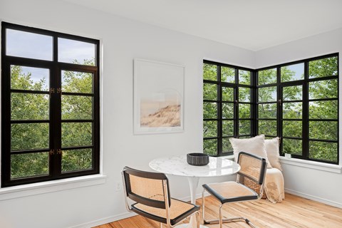 A white table with two chairs is in front of a window with a view of trees.