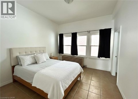 A bedroom with a bed, two pillows, a radiator, and a window with black curtains.
