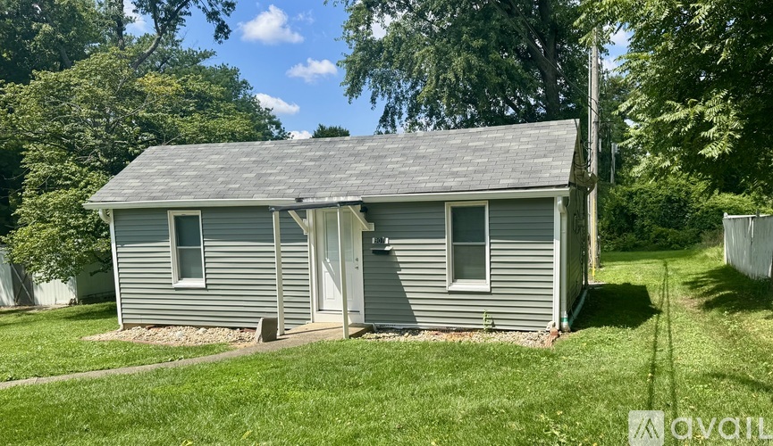 A small house with a grey roof and a white door is surrounded by green grass and trees.