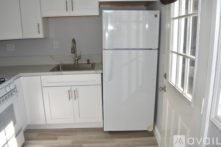 A white refrigerator stands in a kitchen with white cabinets and a stainless steel sink.