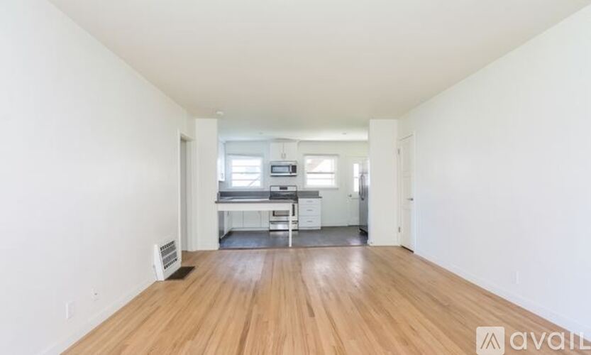 A room with wooden flooring and white walls, leading to a kitchen area.