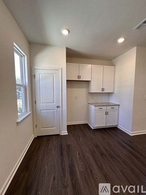 A room with white walls and wooden flooring, featuring a white door, a window, and a kitchenette with white cabinets.
