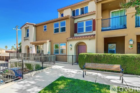 A sunny day at the outdoor patio of a two-story apartment building with a bench and a hedge.