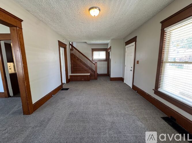 A spacious hallway with a carpeted floor, wooden staircase, and a window with blinds.