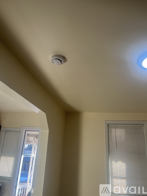 A ceiling with a smoke detector and a window with blinds.