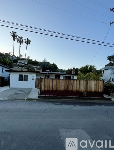 A house with a white exterior and a brown fence is surrounded by a street and palm trees.