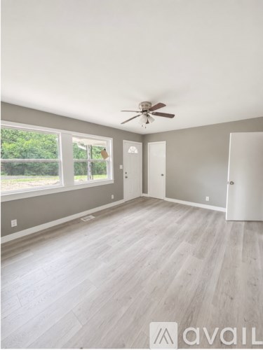 A room with a ceiling fan and light, hardwood floors, and a large window with a view of trees.